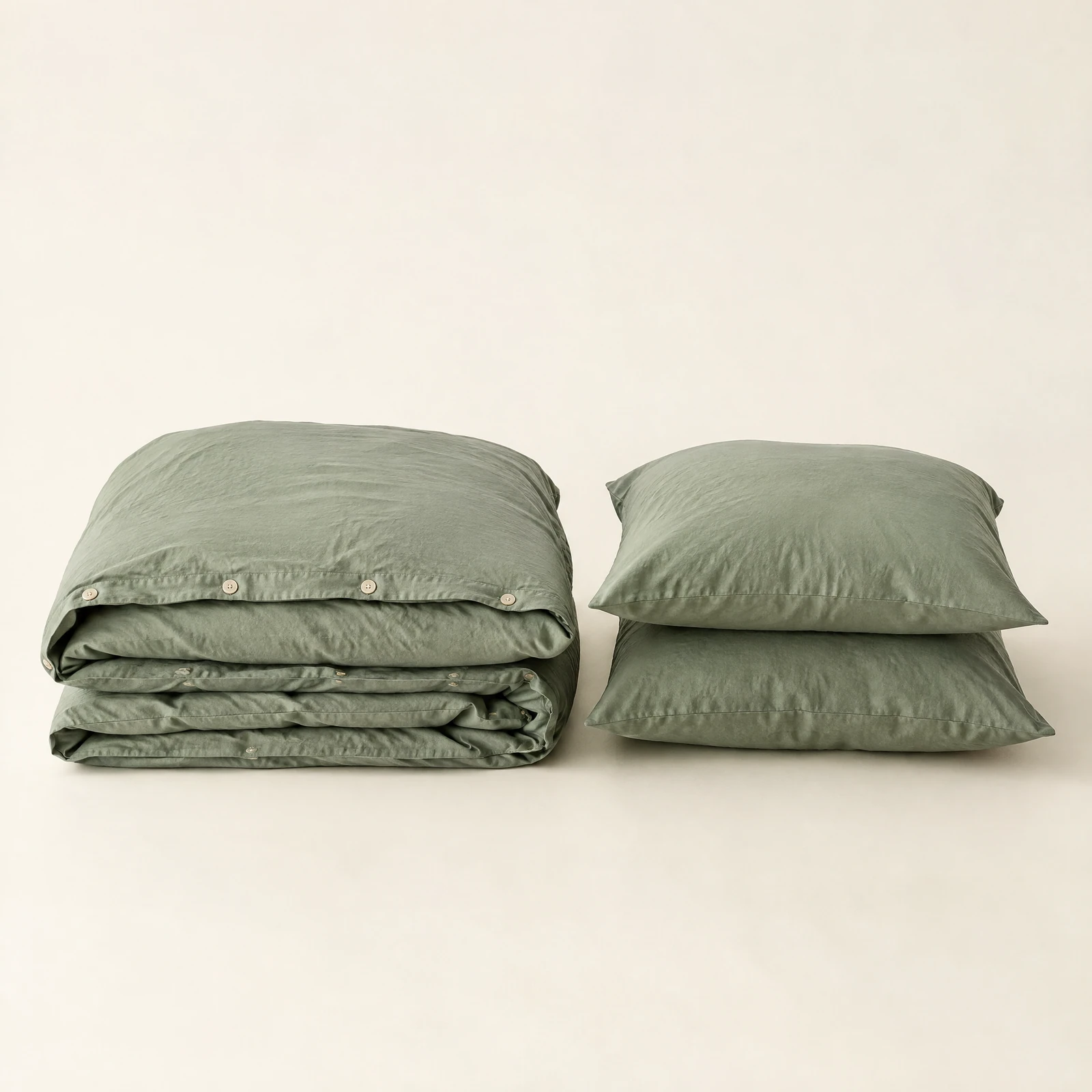 Sage green duvet cover folded beside two matching pillow shams stacked on a cream backdrop